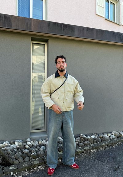 Chaqueta beige con un collar negro, cuatro bolsillos frontales; vaqueros de pierna ancha azul claro; zapatos rojos con acentos blancos; de pie contra una pared gris.