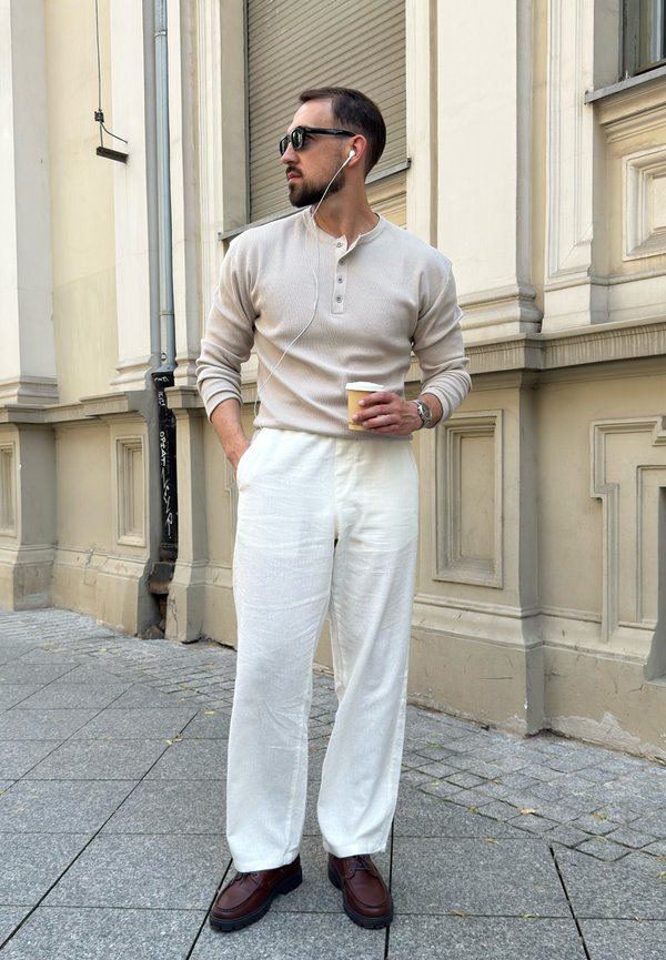 Ein Mann trägt ein beigefarbenes Langarmhemd, weiße Hose und braune Schuhe, steht auf dem Bürgersteig in der Stadt, hält einen Kaffeebecher und trägt eine Sonnenbrille und Kopfhörer.