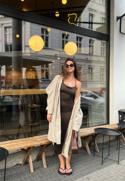 Vestido ajustado marrón con un abrigo trench beige, gafas de sol negras y chanclas. Banco de madera y mesas circulares negras en el fondo.