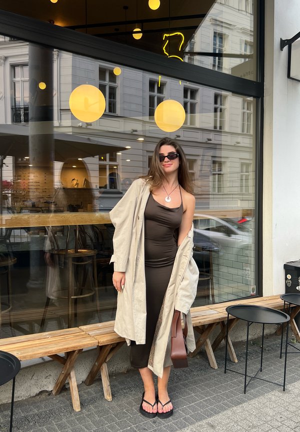 Brązowa sukienka bodycon, beżowy płaszcz trench, czarne okulary przeciwsłoneczne i klapki. Drewniana ławka, czarne stoły i okrągłe żółte lampy w tle.