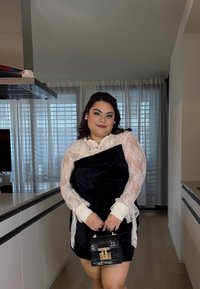 Vestido preto de veludo com mangas de renda e punhos brancos; bolsa preta presa com ferragens douradas; de pé numa sala espaçosa e bem iluminada.