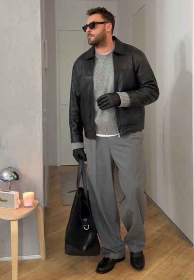 Hombre con chaqueta de cuero negra, suéter gris, gafas de sol, guantes negros, pantalones grises, sosteniendo una bolsa negra en el interior cerca de una mesa con vela y radio.