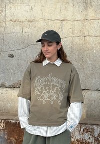 T-shirt à manches courtes vert olive avec un motif en contour blanc, superposé sur une chemise à manches longues blanche à rayures bleues. Une casquette sombre complète le look.