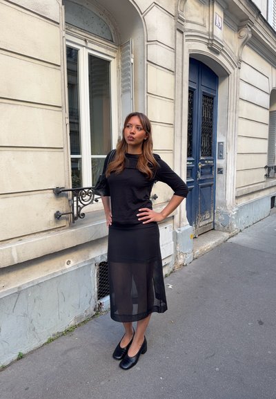 Mujer con top negro y falda negra transparente de pie en la acera junto a un edificio beige con puerta azul y ventana con contraventanas.