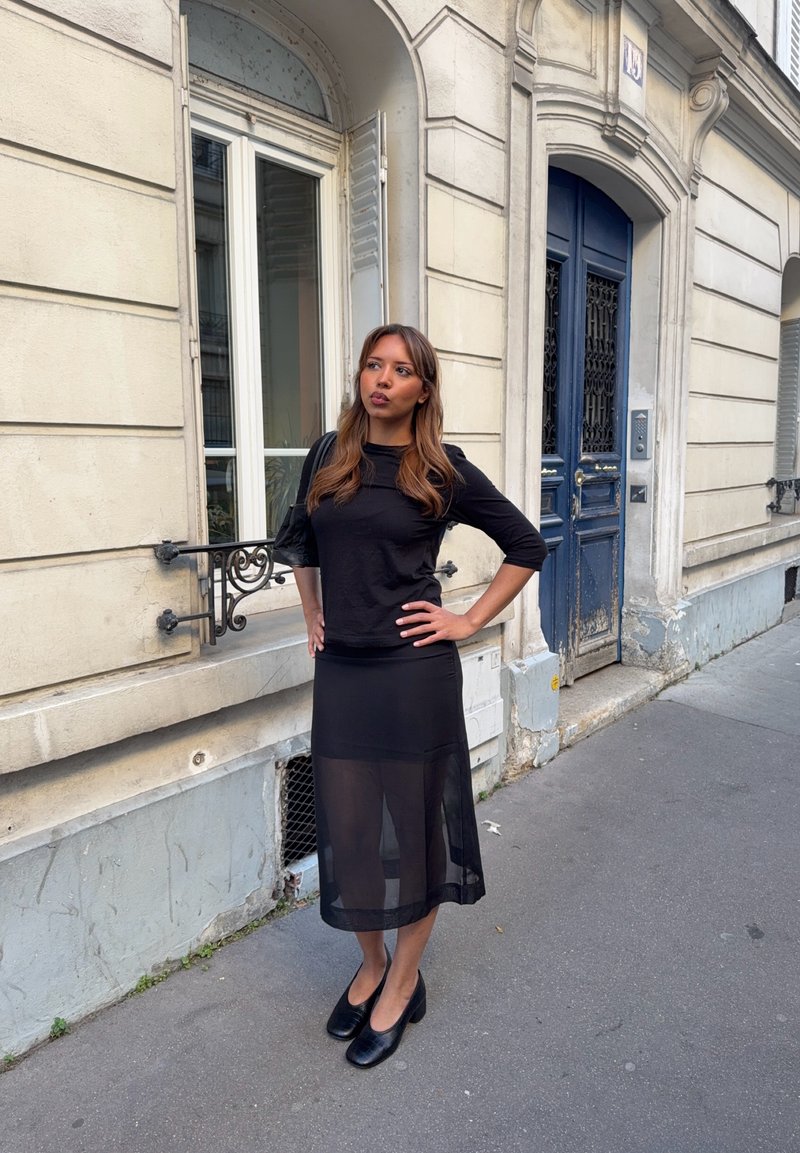 Femme en haut noir et jupe noire transparente debout sur le trottoir près d'un bâtiment beige avec une porte bleue et une fenêtre avec des volets.