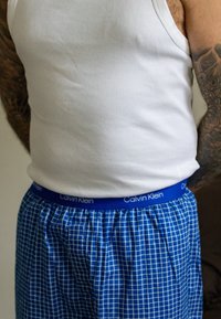 Boxers pour hommes Calvin Klein à motif à carreaux bleus, ceinture élastique avec logo de la marque, associés à un débardeur blanc ajusté.