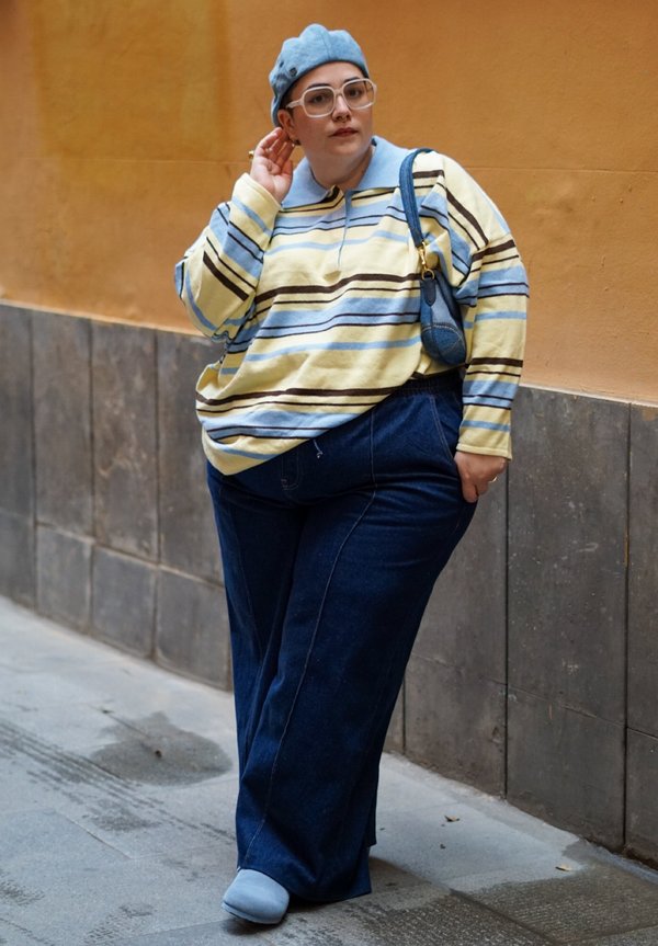 Persona que lleva una boina azul, gafas grandes, jersey a rayas, vaqueros oscuros de pierna ancha y zapatos azules, de pie contra una pared amarilla.