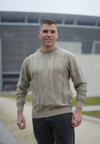 Pull en maille beige avec des lignes verticales texturées, poignets et ourlet côtelés, porté avec un jean gris foncé, sur un fond d'architecture moderne.