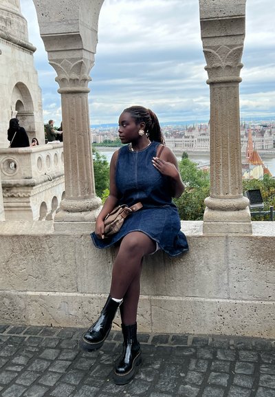 Vestido de mezclilla con un dobladillo acampanado, combinado con botines de plataforma negros y brillantes. Los accesorios incluyen un bolso pequeño y pendientes. Fondo de arco de piedra.