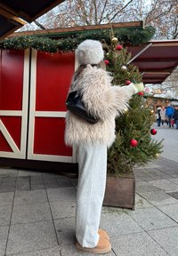 Veste en fourrure beige duveteuse, pantalon de survêtement gris, bottes couleur sable, et un sac à dos noir. Debout à côté d'un sapin de Noël décoré dans un marché.
