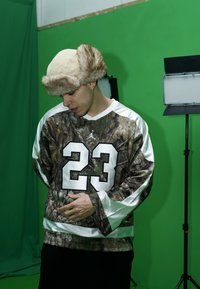 Langermet skjorte med kamuflasjemønster og hvite ermer, med nummer 23. Beige lue med pelskant og svarte bukser, mot en grønn bakgrunn.