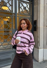 Młoda kobieta z kręconymi włosami, ubrana w różową, w paski koszulę polo oraz brązowe spodnie, trzymająca kubek kawy, stojąca obok dekoracyjnych drzwi metalowo-szklanych.