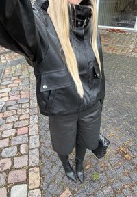 Veste en cuir noir à col haut, associée à un short assorti et des bottes montantes jusqu'aux genoux. Accents métalliques argentés, texture lisse.