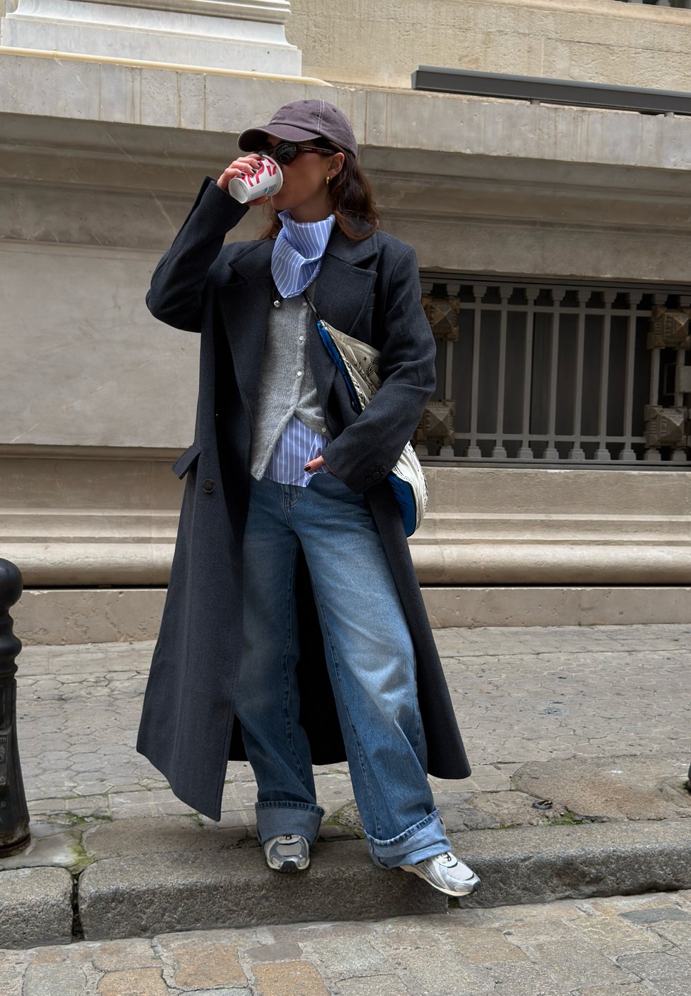 Γυναίκα με σκούρο παλτό, τζιν, καπέλο και αθλητικά παπούτσια πίνει από ένα κουτάκι ενώ στέκεται σε πεζοδρόμιο με κυβόλιθους.