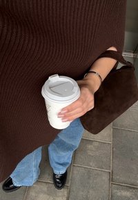 Barna bordázott pulóver, világoskék, bő szárú farmernadrág, fekete cipő, fehér kávésbögrét tartva, sötétbarna velúr táskával.