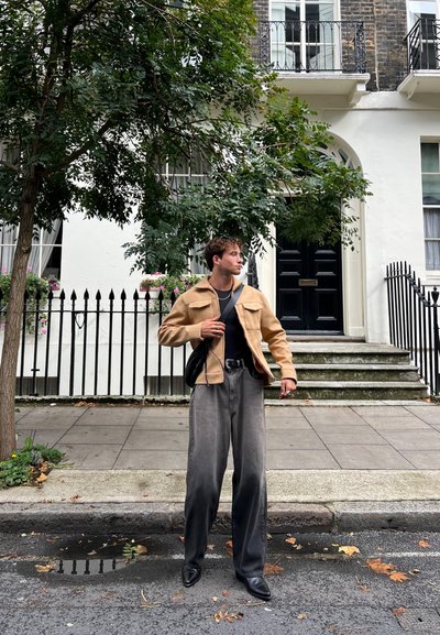 Chaqueta beige con dos bolsillos frontales, top negro y pantalones grises de pierna ancha. Zapatos negros y bolso bandolera. El fondo presenta una puerta negra y vegetación.