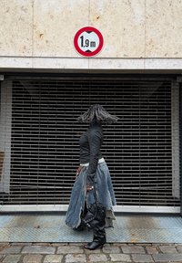 Džínová sukňa s rozstrapkanými okrajmi, čierny dlhý rukáv a čierne čižmy po kolená. Značka označuje výškové obmedzenie 1,9 m nad kovovou mriežkou.