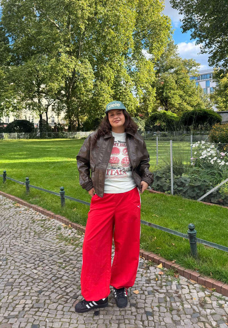 Veste en cuir marron, t-shirt gris avec un motif graphique, pantalon large rouge, baskets noires avec des rayures blanches, casquette de baseball verte portant l'inscription "ciele".