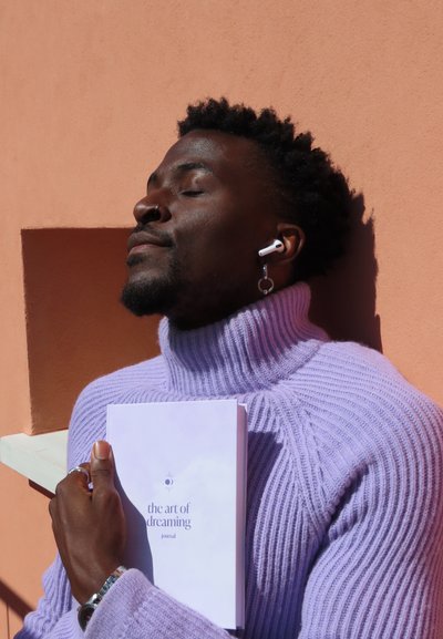 Suéter morado de cuello alto con canalé, sosteniendo un diario blanco titulado "el arte de soñar". Se ven unos auriculares blancos y una pulsera de plata.