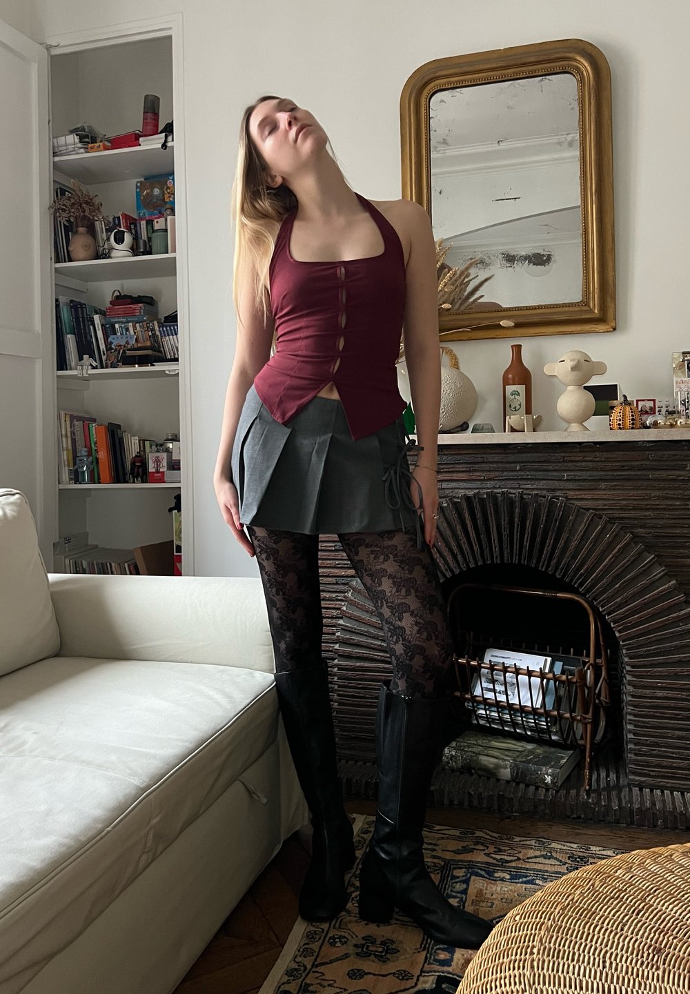 Top halter bordeaux com botões, saia mini plissada cinza, meias pretas de renda e botas altas pretas, ambientado em um aconchegante ambiente interno.