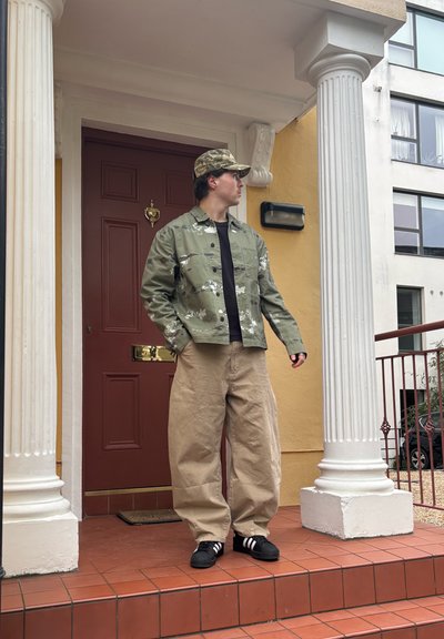 Joven con gorra y chaqueta de camuflaje, pantalones anchos beige y zapatillas negras, de pie en el porche con puerta roja y columnas blancas.