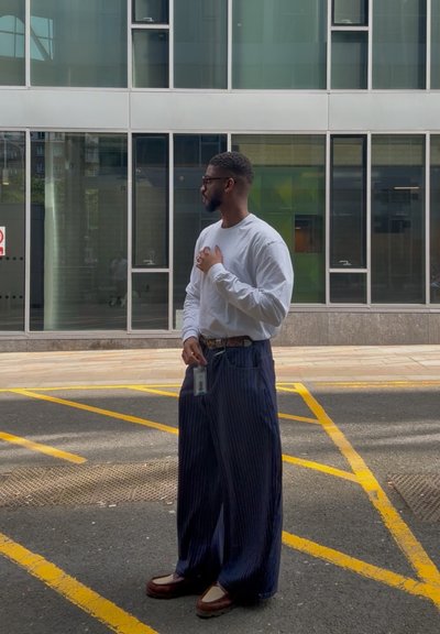 Pantalones navy de rayas finas, holgados y largos, camisa blanca de manga larga, gafas y zapatos marrones. La credencial cuelga de la cinturilla.
