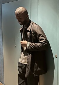 Άνδρας με κοντά μαλλιά και σκουλαρίκι, φοράει σκούρο σακάκι και ριγέ πουλόβερ, στέκεται μπροστά σε ανοιχτό γαλάζιο τοίχο σε εσωτερικό χώρο.