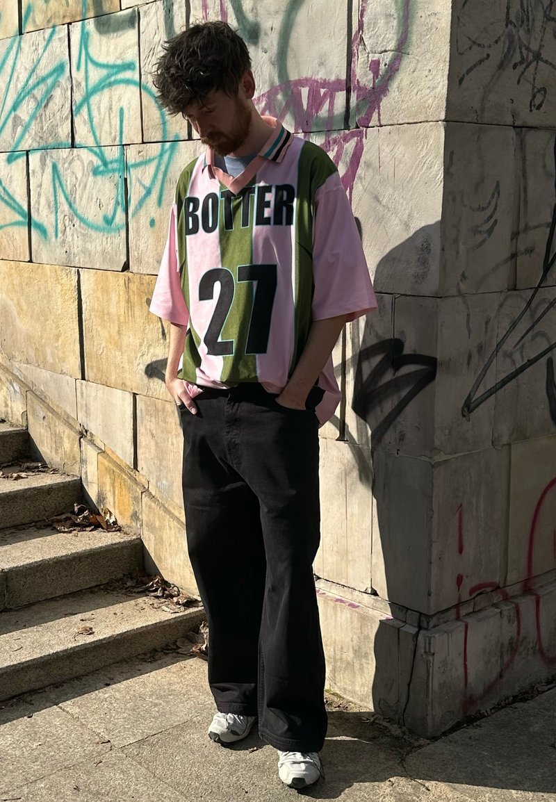 Mann trägt übergroßes rosa-grünes "BOTTER 27"-Shirt, schwarze Hose und weiße Turnschuhe, steht mit den Händen in den Taschen in der Nähe einer Graffiti-Wand.