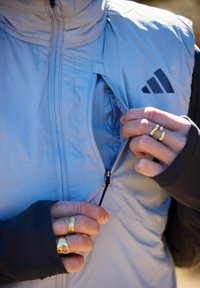 Személy, aki kihúzza a világoskék Adidas dzseki zsebének cipzárját, láthatóvá téve a steppelt bélés egy részét, mindkét kezén aranygyűrűkkel.