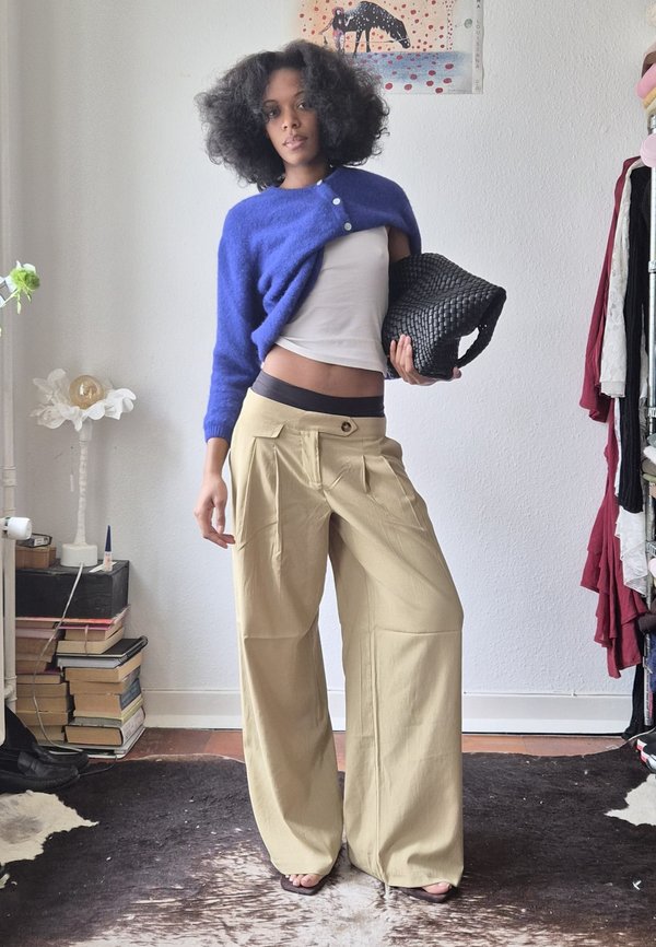 Woman with curly hair wearing a blue cropped sweater, beige wide-leg pants, holding a black woven bag, standing in a room with books and clothes.