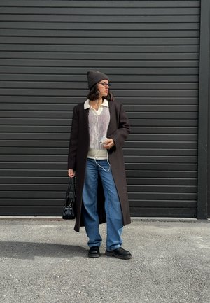 Mujer con abrigo largo marrón, suéter beige, jeans azules, zapatos negros y gorro de lana sostiene un bolso negro y un teléfono con auriculares al aire libre.