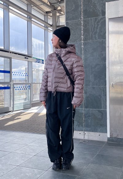 Chaqueta acolchada en malva con capucha, combinada con pantalones deportivos negros con rayas blancas. Un gorro negro y unas zapatillas oscuras completan el look.