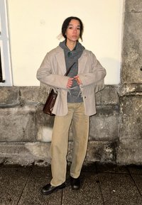 Γκρι φραγκογελέκο πάνω από μια γκρι φούτερ, καμηλό παντελόνι και μαύρες δερμάτινες μπότες. Κρατά μια καφέ υφασμάτινη τσάντα. Ουδέτερο φόντο, χαμηλό φωτισμό.