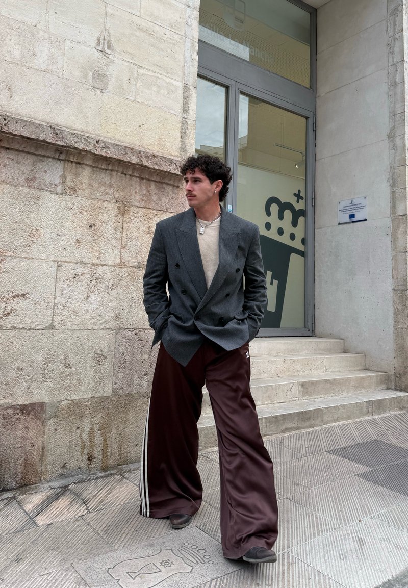 Homme aux cheveux bouclés portant un blazer gris croisé, une chemise beige et un pantalon marron large, debout dans une rue pavée près de l'entrée d'un bâtiment.