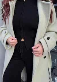 Gai pelēks vilnas mētelis ar lielām melnām pogām, uzvilkts virs pieguļošas melnas ribotas kapuces augšdaļas un melnām biksēm ar auklu. Gluda tekstūra un slāņošana.