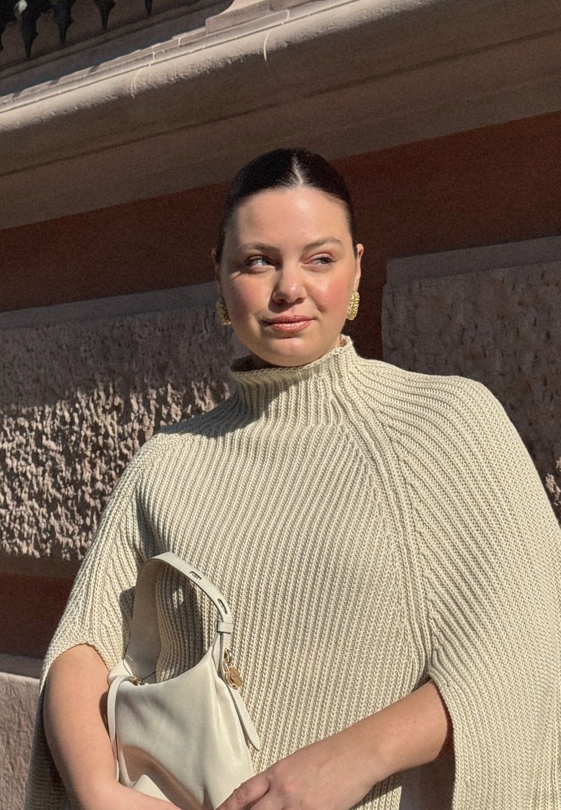 Mujer de cabello oscuro con un poncho beige de punto sostiene un bolso blanco, de pie al aire libre contra una pared texturizada bajo la luz brillante del sol.