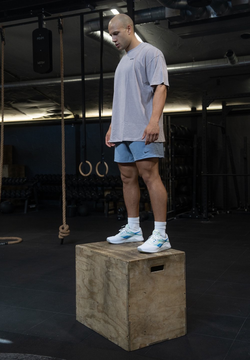 Caixa de madeira numa academia com uma pessoa em pé em cima dela. A pessoa veste uma t-shirt cinza clara, calções azuis e sapatilhas brancas com detalhes azuis.