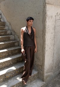 Brązowy top na ramiączkach z gładkiego materiału, zestawiony z luźnymi brązowymi spodniami. Teksturowana brązowa kopertówka w dłoni, stoi na tle teksturowanej ściany.