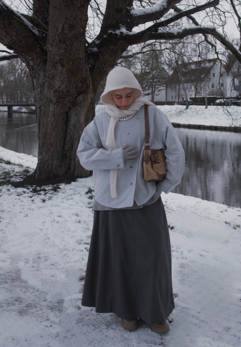 Femme portant une veste blanche à capuche, une écharpe et une jupe grise, debout à côté d'un grand arbre près d'une rive enneigée en hiver.