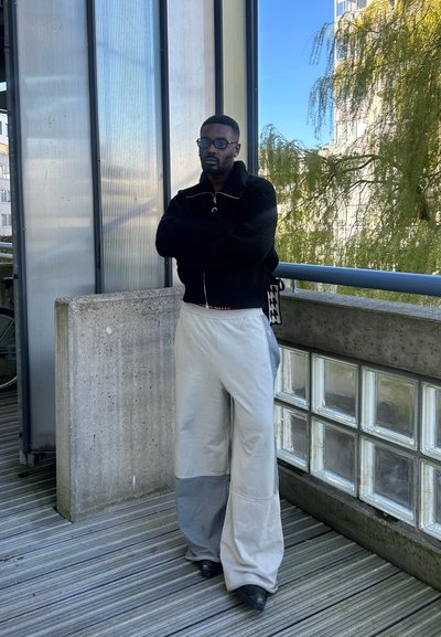 Chaqueta de punto negra con cremallera, pantalones grises holgados con una sección inferior gris oscuro, y zapatos negros. Fondo de pared de vidrio y hormigón texturizado.