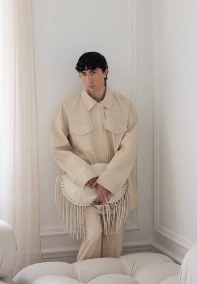 Persona joven con cabello rizado oscuro lleva una chaqueta y pantalones beige oversize, sostiene un bolso crema con flecos, de pie en una esquina luminosa de una habitación.