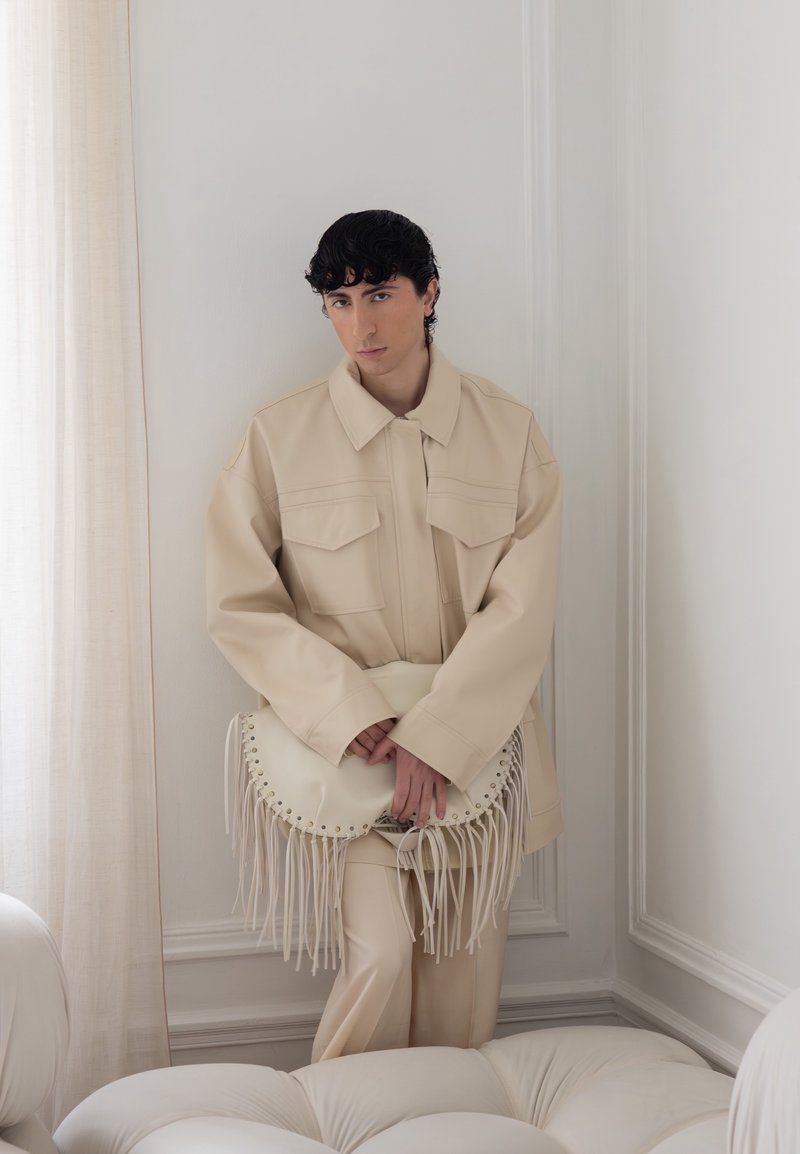 Young person with dark curly hair wears oversized beige jacket and pants, holding a cream-fringed handbag, standing in a bright room corner.