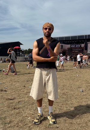 Schwarzes, geripptes, ärmelloses Top, beige Shorts, schwarz-gelbe Sneakers und Sonnenbrille. Außeneinstellun mit einer Menschenmenge im Hintergrund.