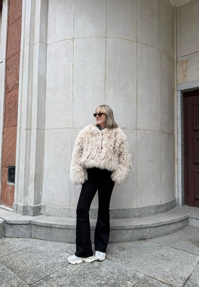 Chaqueta de peluche en beige claro, con una textura esponjosa y frente abierto. Combinada con pantalones de campana negros y zapatillas grises. De pie contra una pared de piedra.