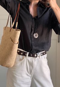 Zwarte overhemd, witte spijkerbroek, bruine riem met zilveren accenten en een geweven tan tote bag. Cirkelvormige hangeren als een opvallend detail.