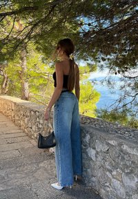 Light blue high-waisted jeans with wide legs, a black crop top, white pointed shoes, and a small black handbag. A flower in the hair. Background of a stone path.