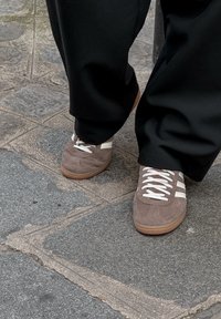Brune ruskinds sneakers med hvide snørebånd og tre hvide striber, parret med sorte vide bukser, på en struktureret stenoverflade.