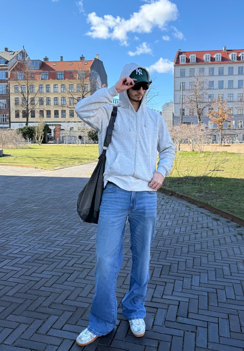 Homme en sweat à capuche gris, jean bleu, baskets blanches, lunettes de soleil et casquette verte ajuste la sangle de sa casquette en se tenant sur une place pavée avec des bâtiments en arrière-plan.
