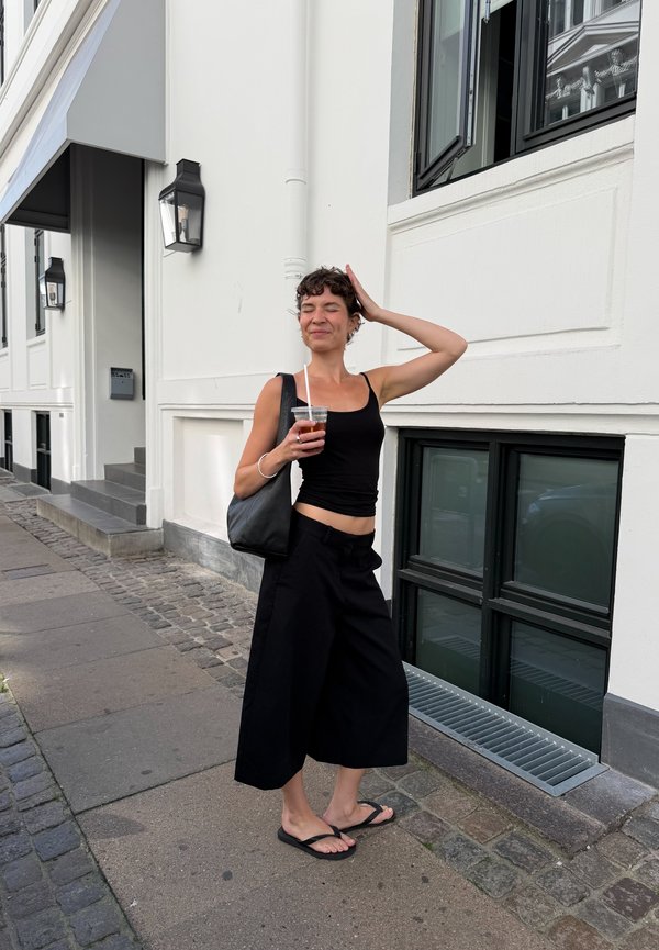 Jeune femme en haut noir et pantalon large tenant un verre, debout détendue sur un trottoir en ville près d'un bâtiment blanc avec des fenêtres ouvertes.