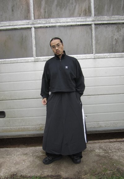 Sudadera negra con cremallera y un corte holgado, combinada con una falda larga negra con rayas laterales, y zapatos negros con suelas texturizadas.